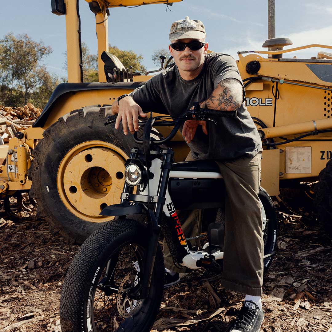 A man wearing dark colored pants, a dark colored t-shirt and a light colored hat sits on the SUPER73-M1D SE in Baja. Behind him, a large yellow piece of farming equipment is visible.