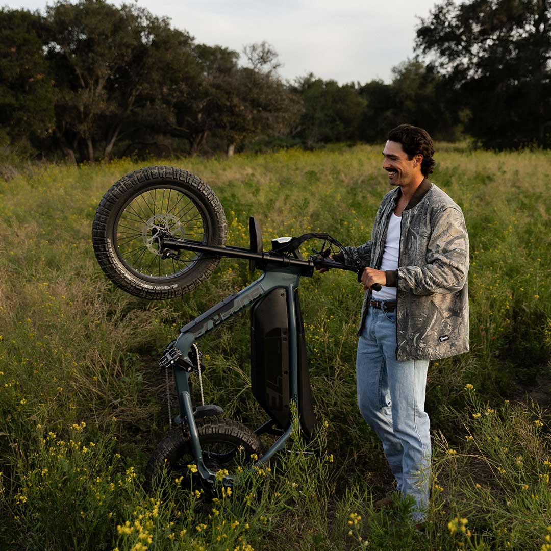 A man wearing light colored jeans, a white top and a light colored jacket stands in a green field while holding the handlebars of a SUPER73-M1D. The ebike is vertical and resting only on its back tire with the front tire facing up.