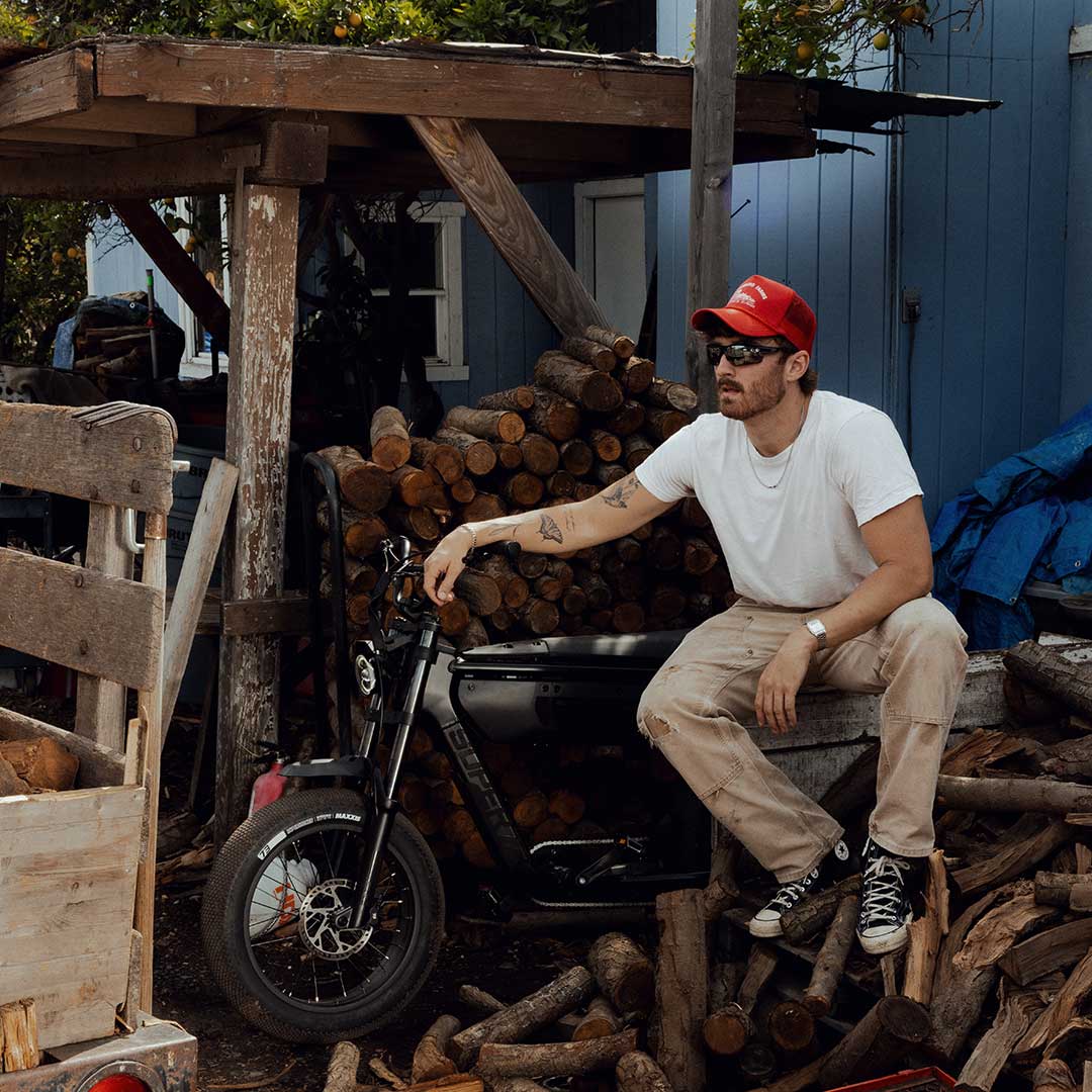 A man wearing light colored pants, a white t-shirt and a red trucker hat sits on a large pile of chopped wood with his right arm resting on the handlebars of a SUPER73-B1G SE in Blackout.