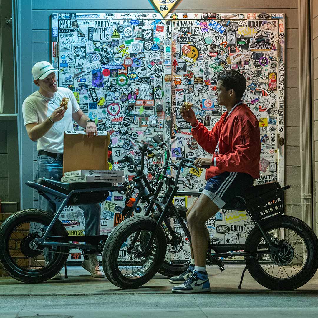two young adults eating pizza in front of a door filled with stickers on their ebikes