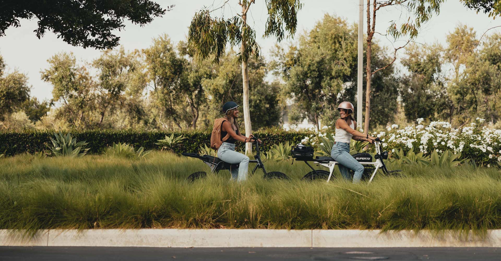 two friends riding their SUPER73s through the park while wearing helmets