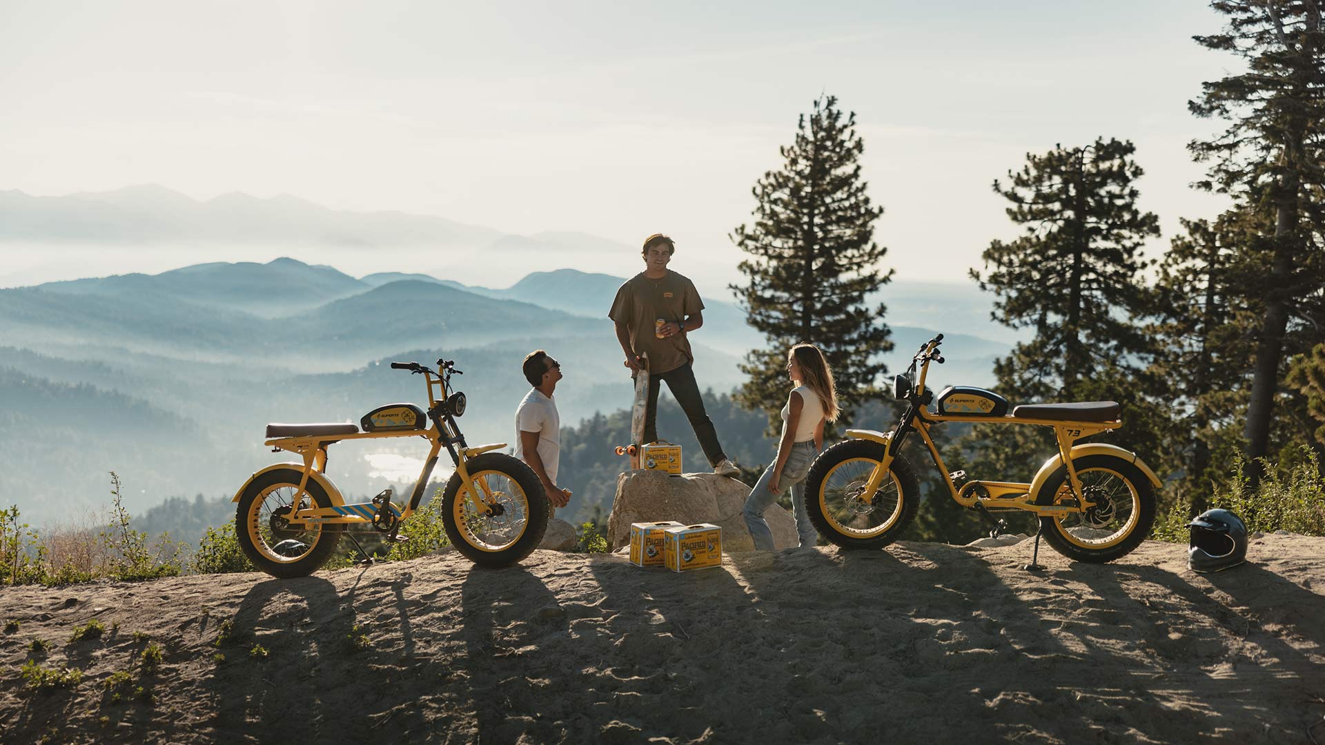 Two men and one woman are standing near a cliff overlooking trees and mountains with two custom Pacifico x SUPER73-S2 bikes and some Pacifico beer.
