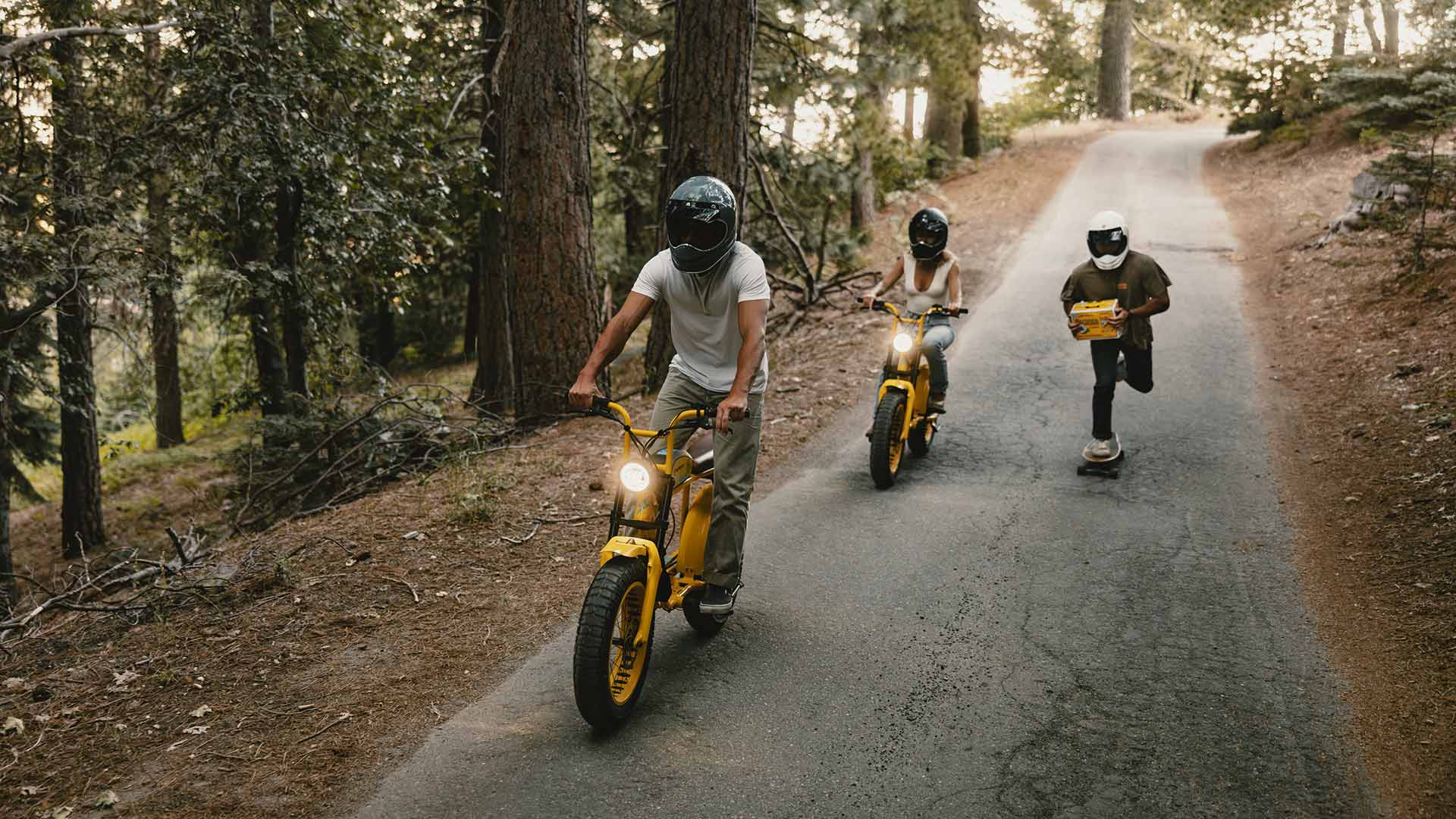 A man and woman riding custom Pacifico x SUPER73-S2 bikes along a path while another man rides a skateboard carrying a case of Pacifico beer.