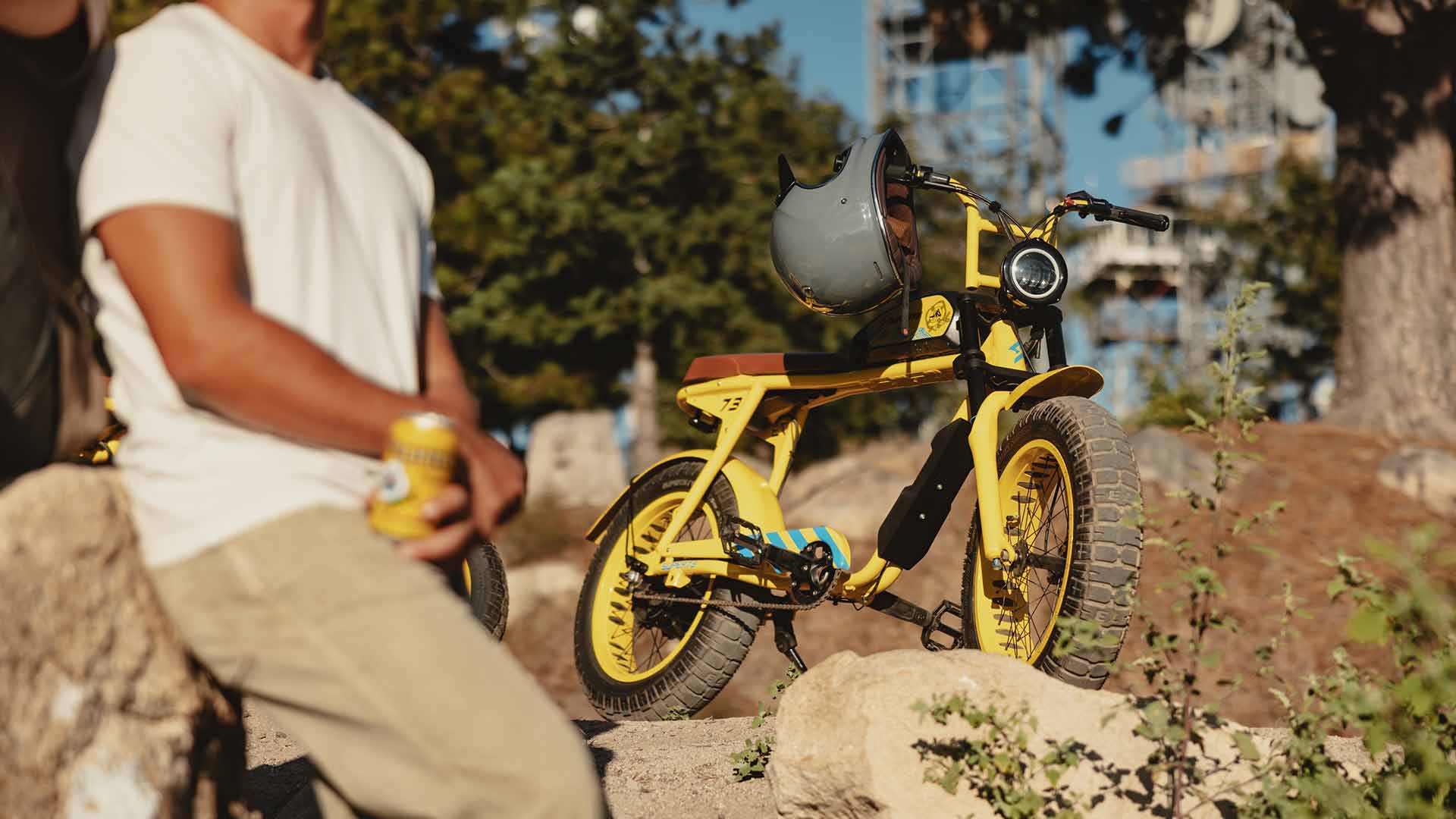 A man leans against a rock holding a can of Pacifico beer with a custom Pacifico x SUPER73-S2 bike stands in the background.