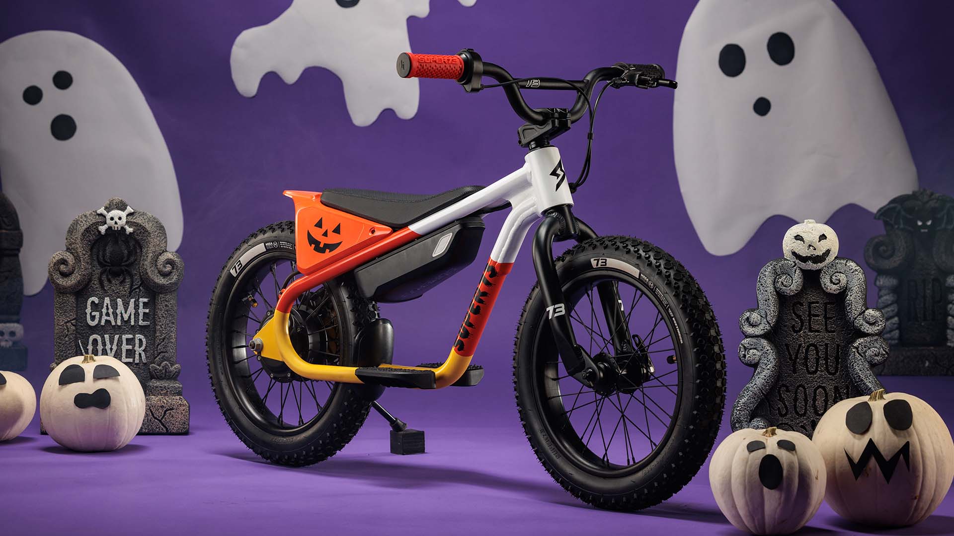 The SUPER73-K1D Candy Corn Cruise on a purple backdrop with pumpkins and ghosts in the background.