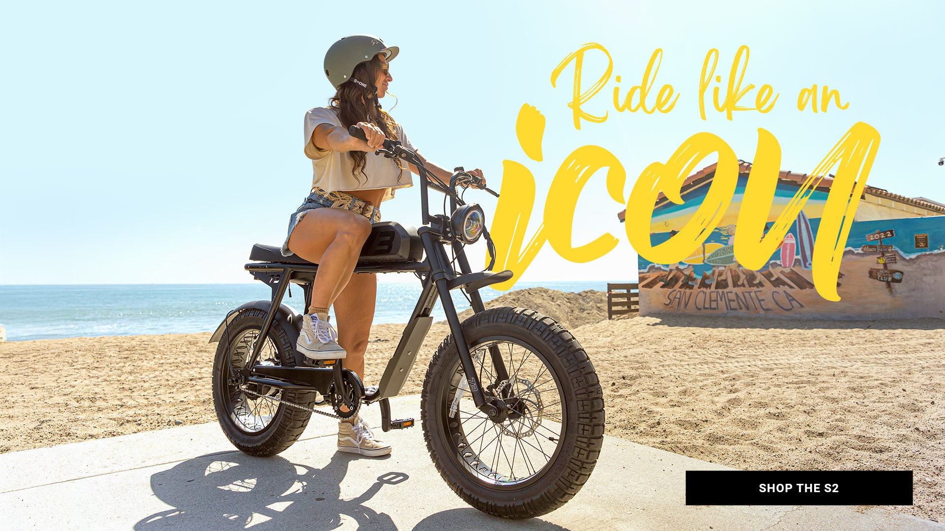 A woman wearing a helmet sits on a SUPER73-S2 Laguna ebike on a concrete surface at the beach.