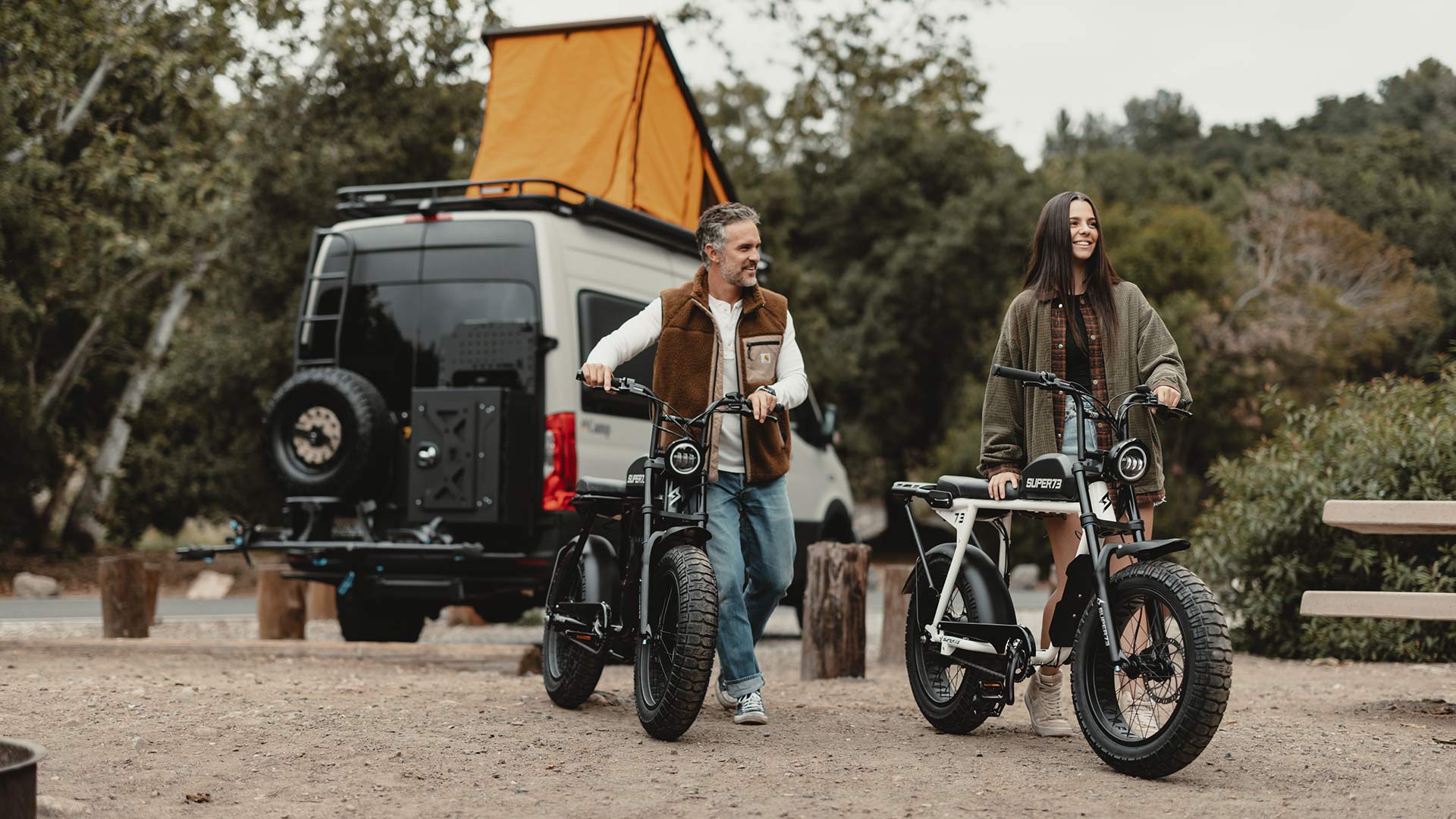 A man and a woman walking their SUPER73-S2 bikes with a utility vehicle in the background.