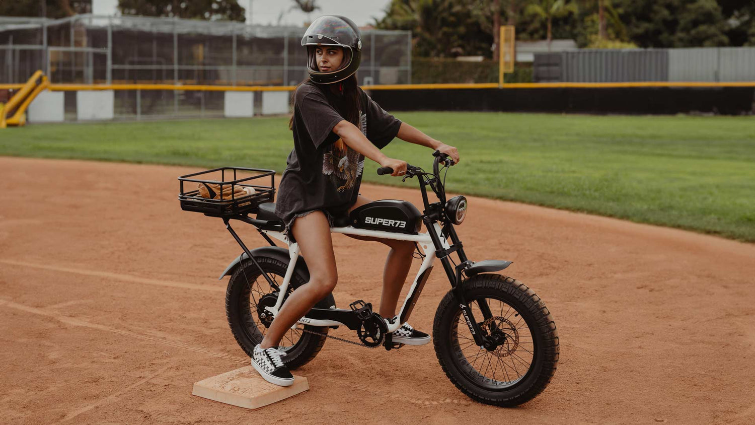 Image of a female rider wearing a helmet and sitting on a SUPER73-S2 ebike on a baseball field.