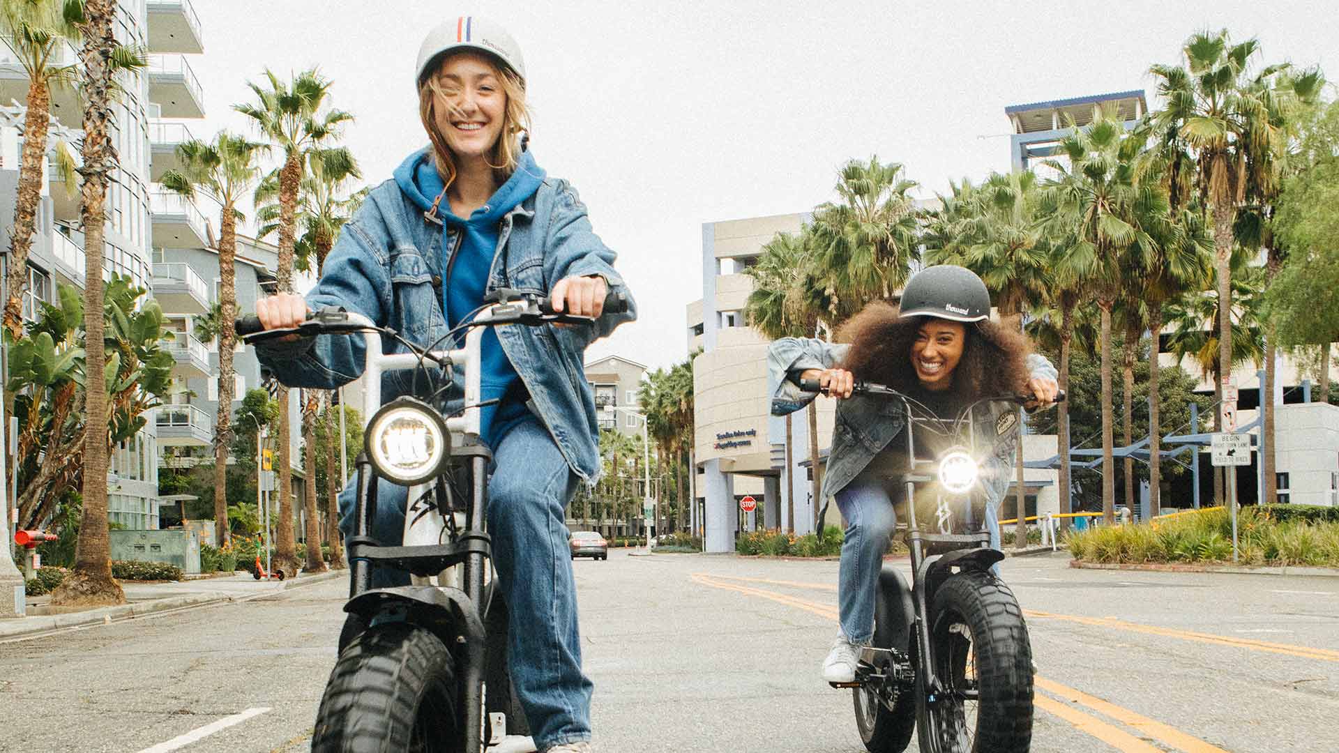 Two female SUPER73 electric bike owners ride down the street together smiling.