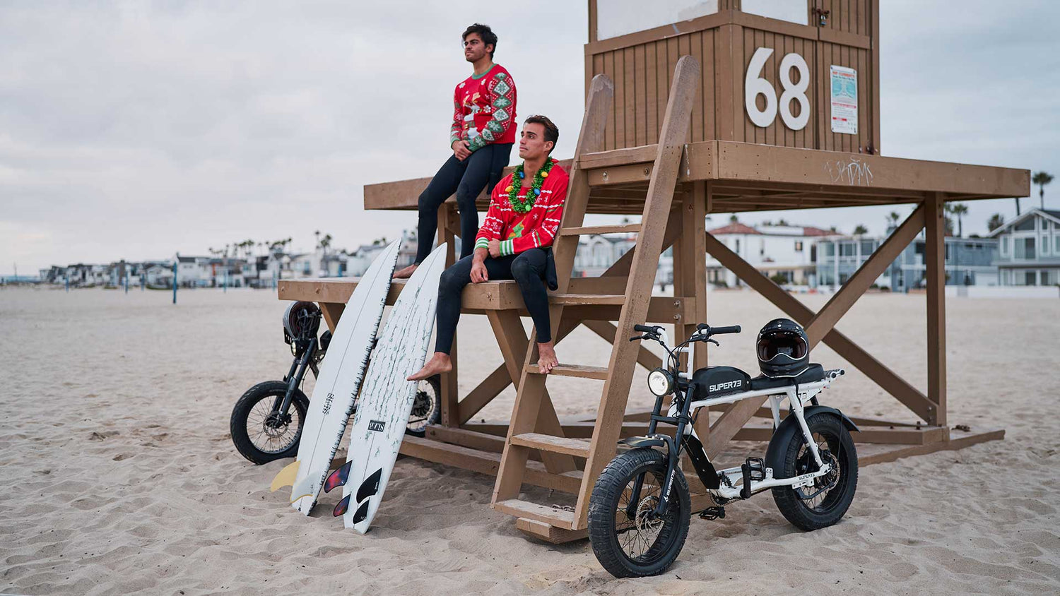 Two men sit on a lifeguard tower in holiday sweaters surrounded by two SUPER73 electric bikes and surfboards.