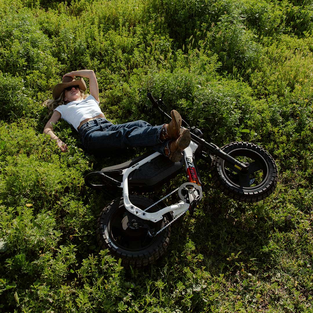 A woman wearing dark jeans, a white-tshirt and a brown cowboy hat is laying on her back in a green field. In front of her, a SUPER73-MZFT SE lays on it's side with her legs propped up on the seat.