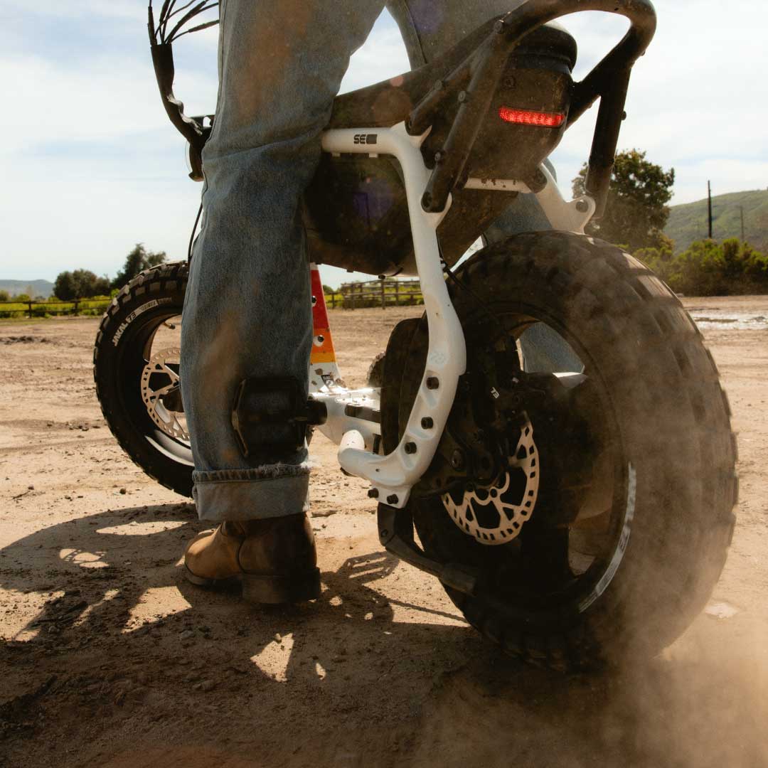 A person wearing jeans and boots stands stradling a SUPER73-MZFT SE in Baja on a dirt path.