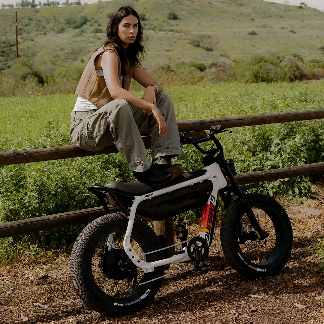 A woman in light colored pants and a light top underneath a tan vest sits on a wooden fence running along a green field. In front of her, a SUPER73-M1D SE is leaning against the fence and the woman is resting her feet atop it.