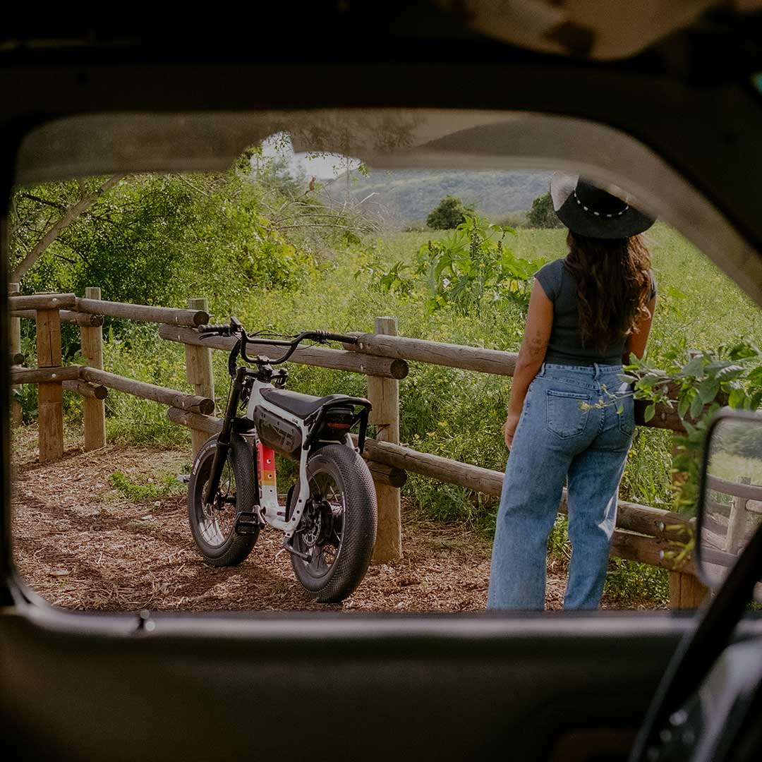 A woman wearing jeans, a black t-shirt and a black cowboy hat stands outside the cab of a truck leaning against a wooden fence. A few feet in front of her, a SUPER73-M1D SE in Baja is parked and resting against the fence with a green field visible in the background.
