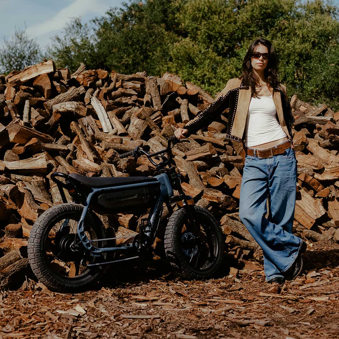 A woman wearing jeans, a white top and a tan and black jacket faces the camera with her right arm outstretched and resting on the handlebars of a SUPER73-M1D. Behind her, a large pile of wooden logs and a tree are visible.