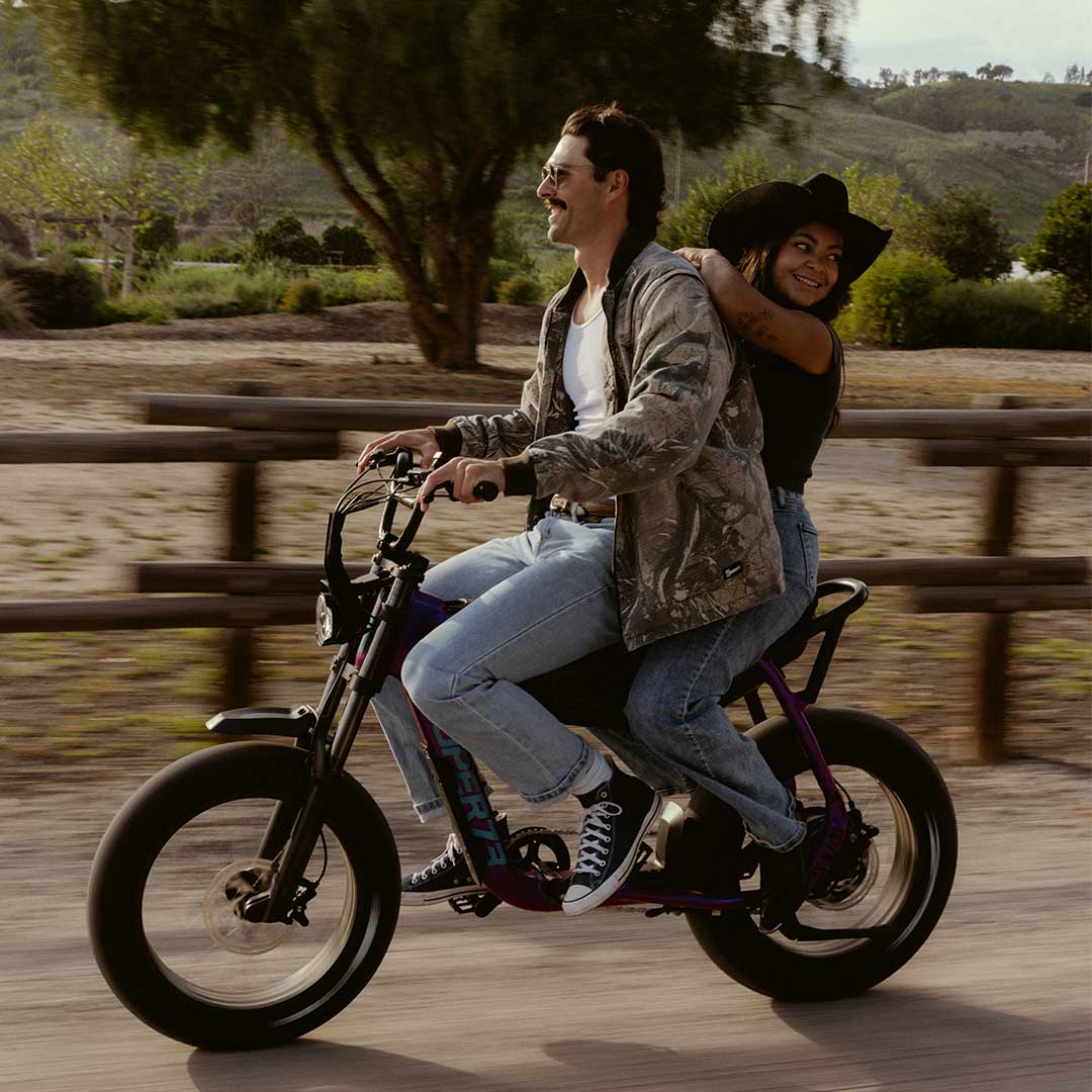 A man wearing jeans, a light colored top and a light colored camo print jacket rides a SUPER73-B1G-SE in Cullenberry in front of a wooden fence. A woman wearing jeans and a black t-shirt sits on the back of the bike, smiling. 