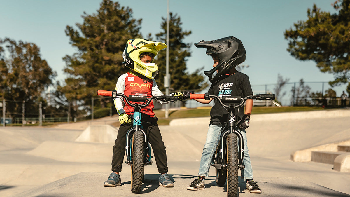 Two young children on SUPER73-K1D ebikes wearing full face helmets and talking