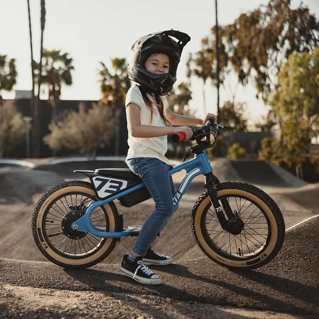 Young rider on her blue SUPER73 K1D kid's e bike on a paved pump track looking at the camera smiling with her helmet on