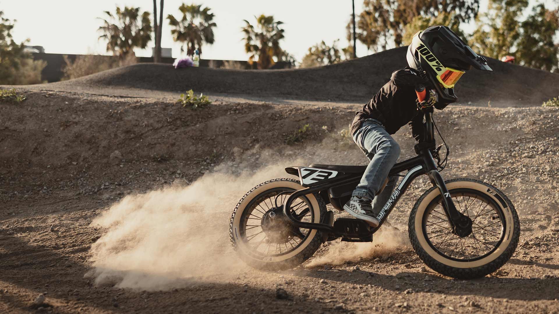 Young boy riding a blue SUPER73-K1D in a helmet on a dirt track kicking up dust