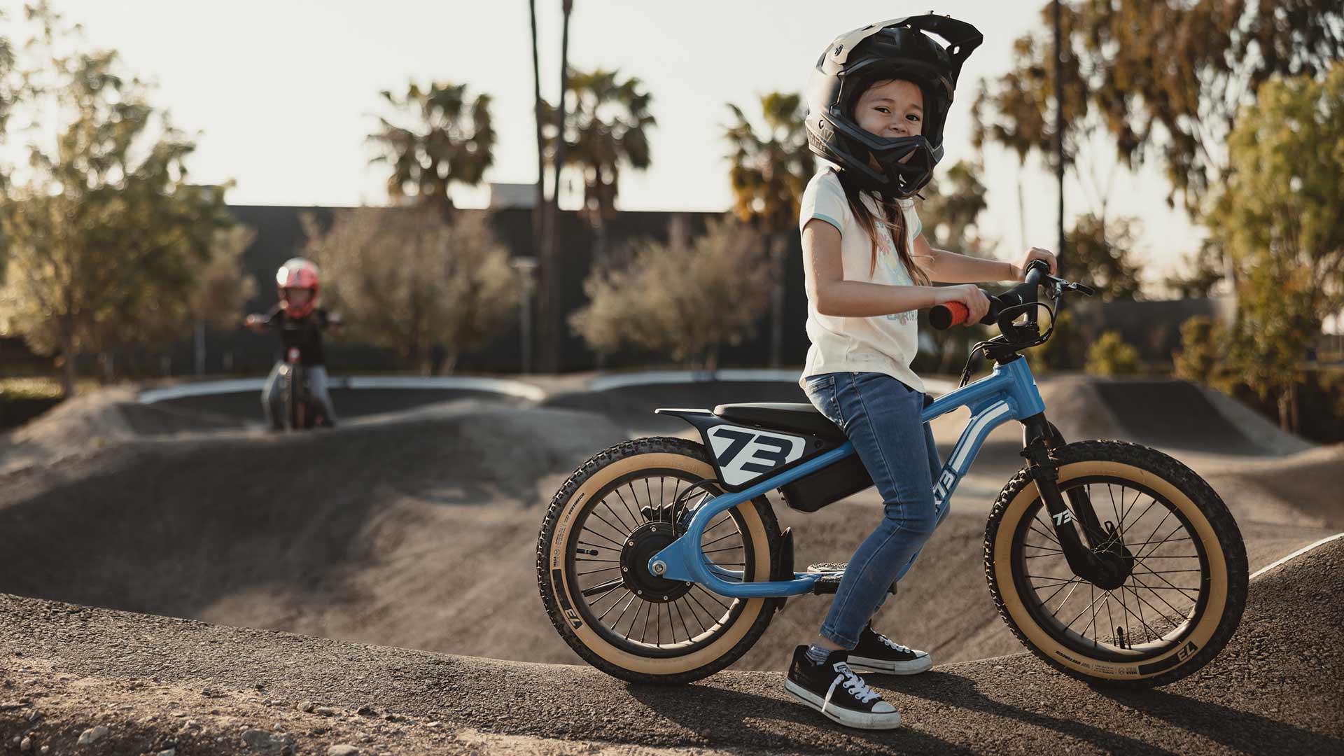 Young girl sitting on a blue SUPER73-K1D in a helmet on a dirt track 