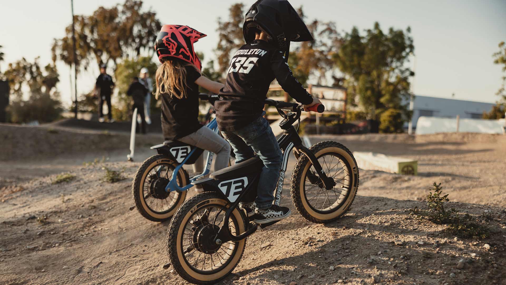 Two young kinds riding the SUPER73-K1D side-by-side in helmets on a dirt track 