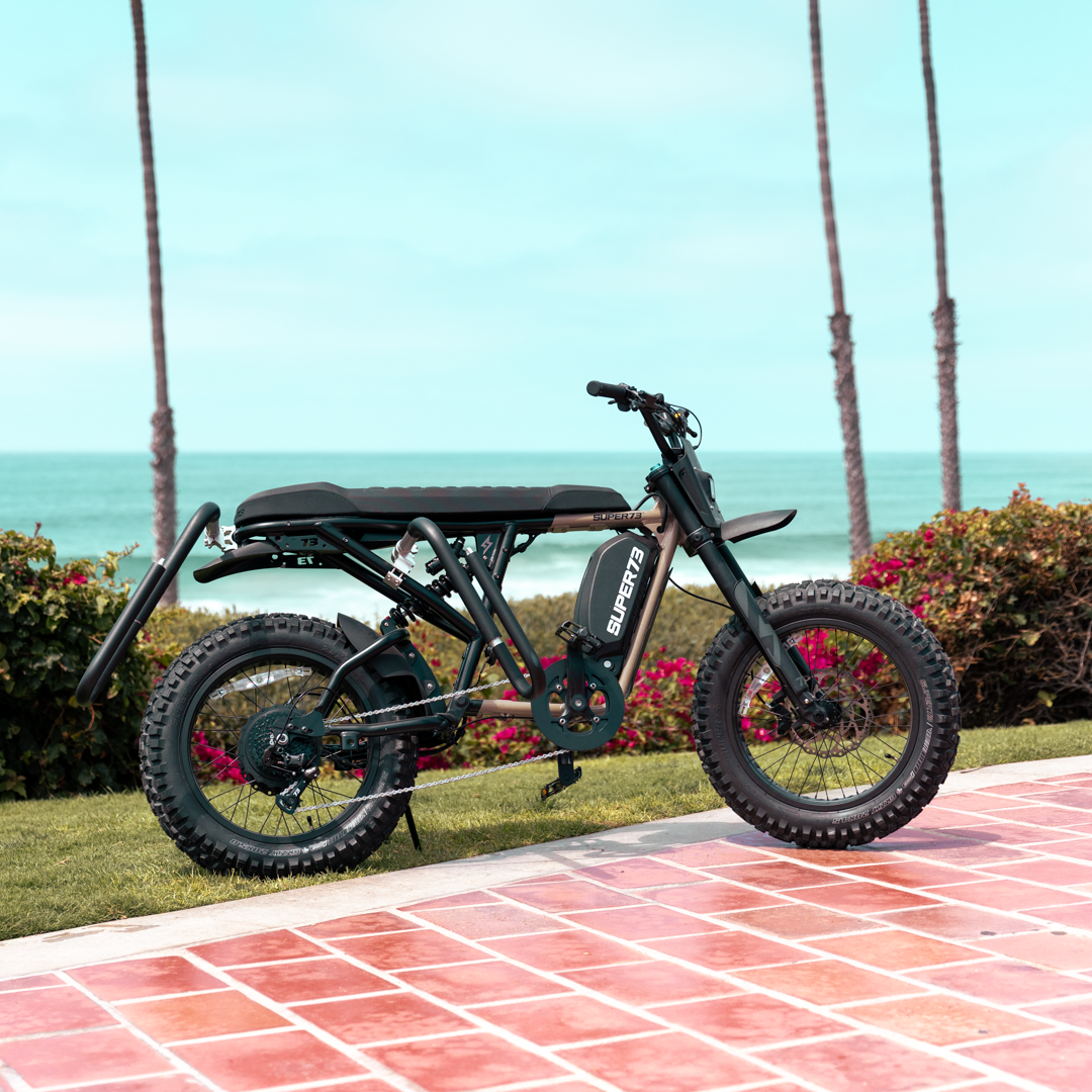 A SUPER73-R Adventure Series sits parked on a grassy area near the beach. A MBB Dual Sport Surf Rack is installed on the bike.