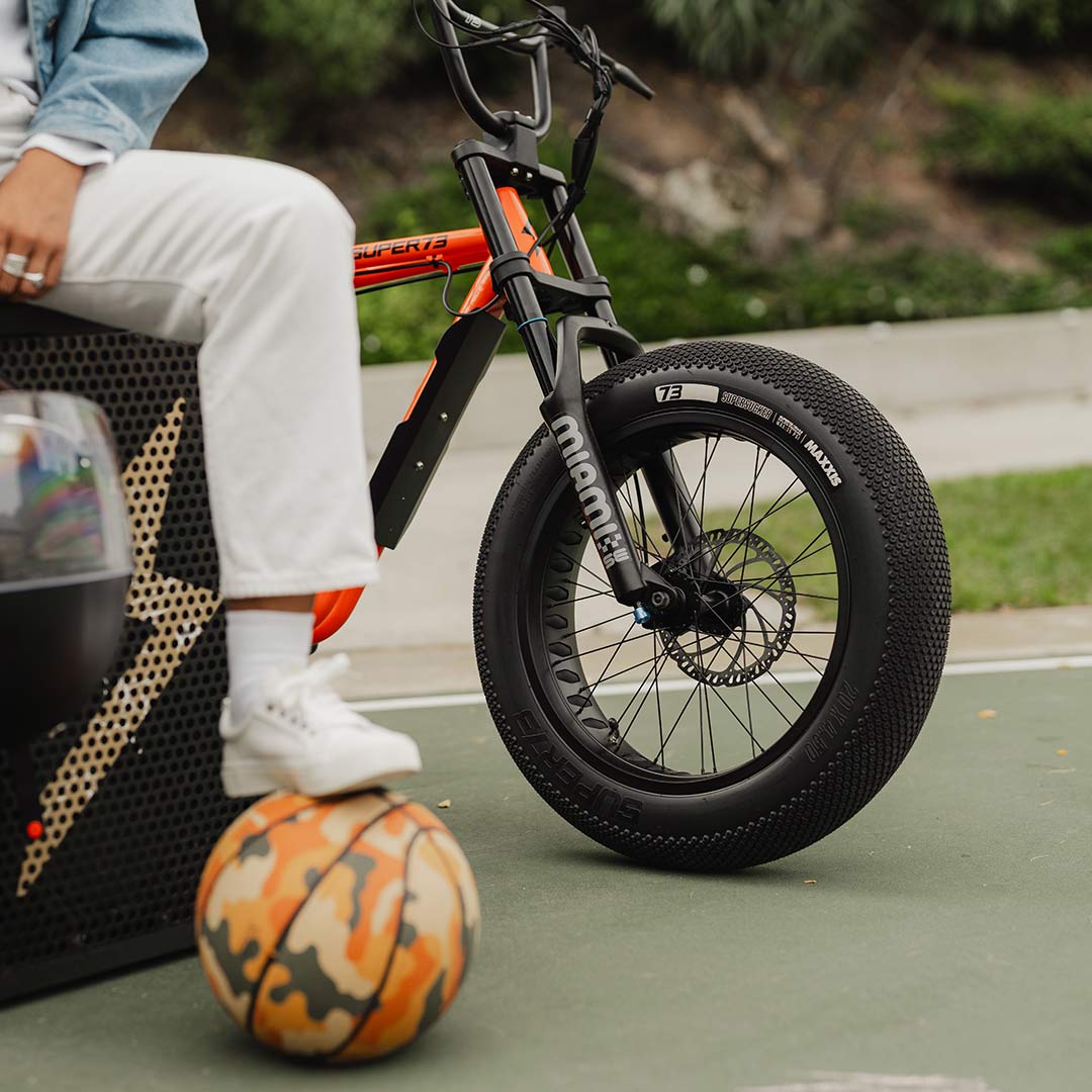 A SUPER73-Z Miami SE bike in Astro Orange next to a person with their foot on a basketball.