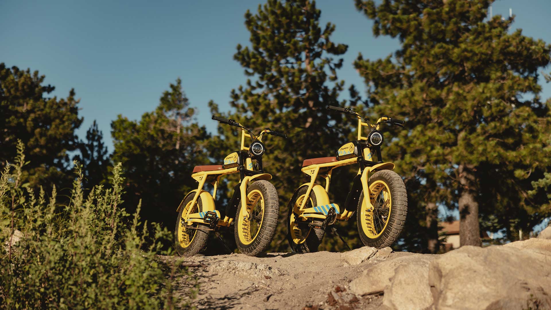 Two custom Pacifico x SUPER73-S2 bikes sit atop a cliff surrounded by trees.