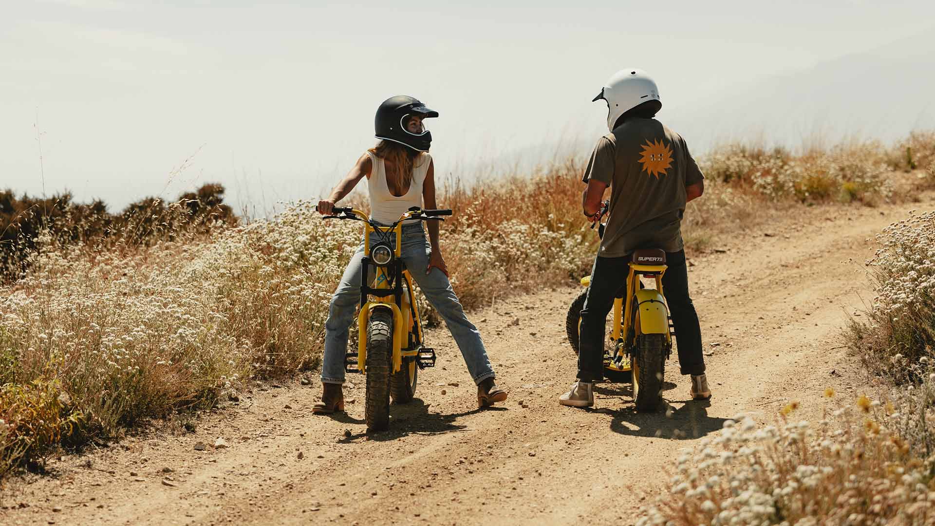 A man and woman facing each other while sitting on their custom Pacifico x SUPER73-S2 bikes on a dirt trail.