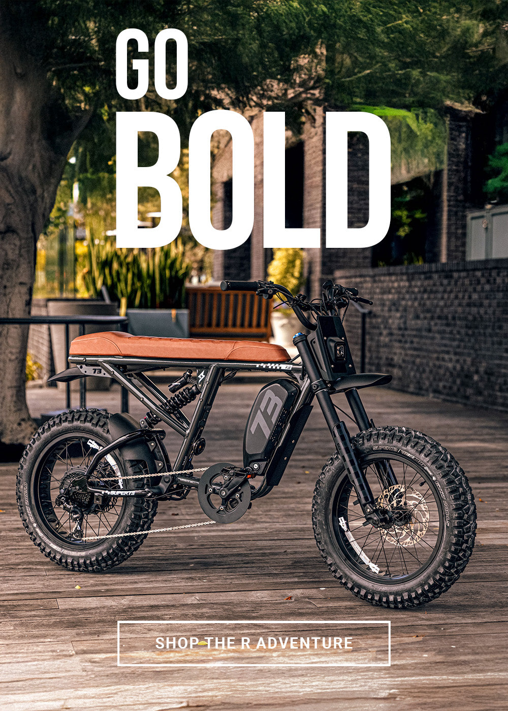 Lifestyle image of the SUPER73-R Adventure SE ebike in Palladium. The bike is parked on a wooden surface with a brick building and plants in the background.