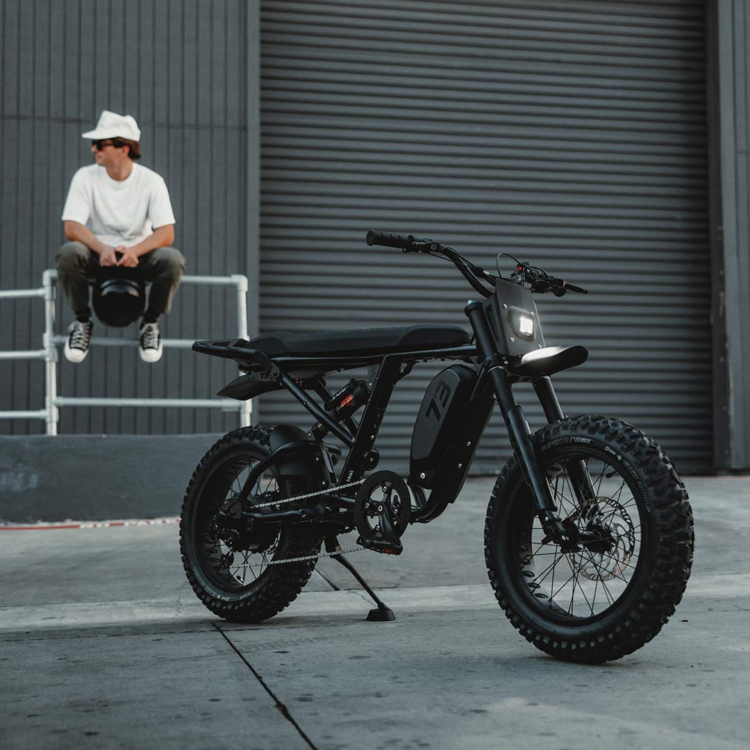 Image of a SUPER73-R Blackout SE bike in the foreground while a man sits on a fence in the background.