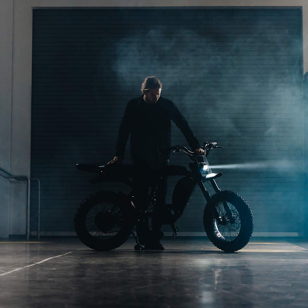A man stands in front of a SUPER73-R Blackout SE bike in a warehouse.