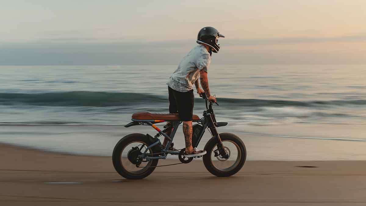 A young man standing on the pedal of a SUPER73 R-Series ebike while riding on the beach