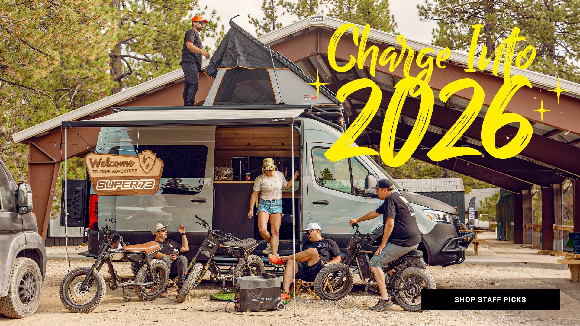 A group of people sit next to a camper van with several ebikes in the foreground. Text overlay reads "Charge into 2026" | Click to shop staff picks.