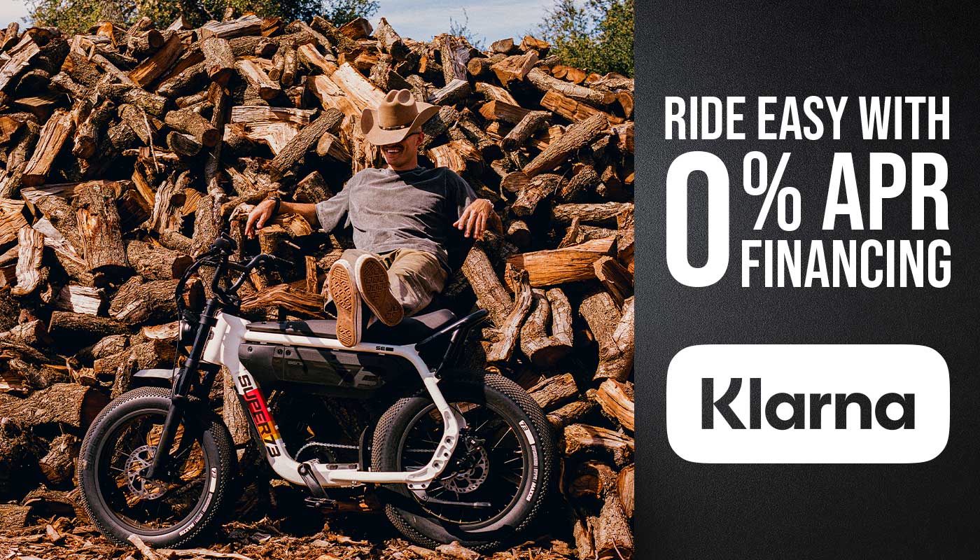 A man sits atop a large pile of wooden logs with his legs and feet resting on the seat of a SUPER73-M1D. Text on the right-hand side of the image reads "Ride easy with 0% APR financing from Klarna"