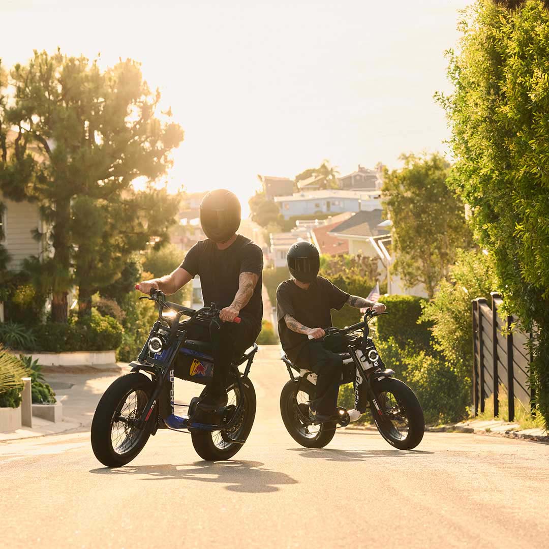 Two men wearing dark helmets riding the SUPER73 x Oracle Red Bull Racing M1D SE ebikes through a neighborhood.