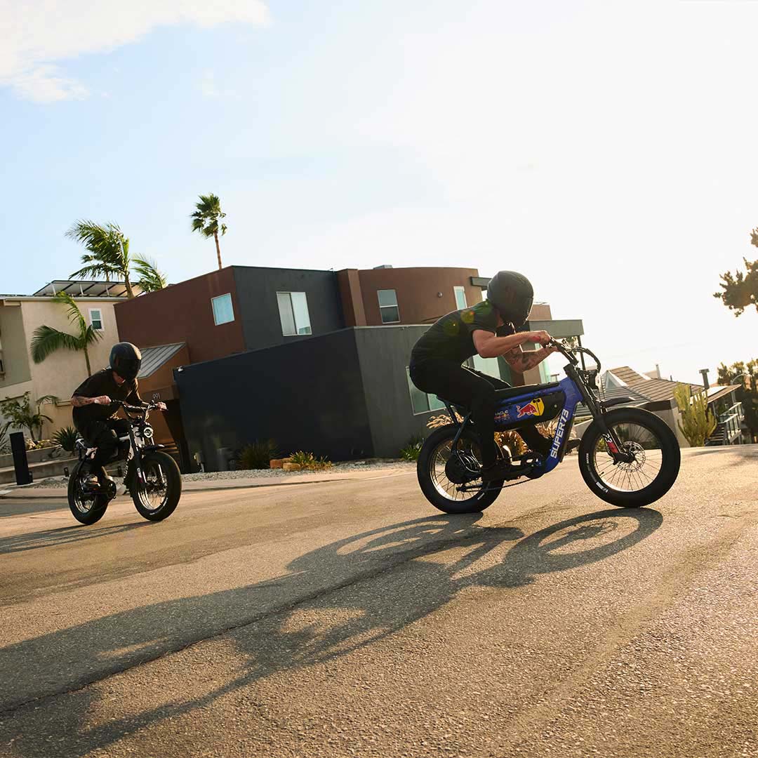 Two men wearing dark helmets riding the SUPER73 x Oracle Red Bull Racing M1D SE ebikes on a paved road with houses in the background.