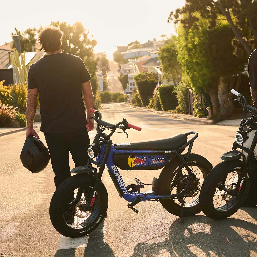 A man stands with his back to the camera holding a dark helmet next to the SUPER73 x Oracle Red Bull Racing M1D SE ebike in Nightfall Blue