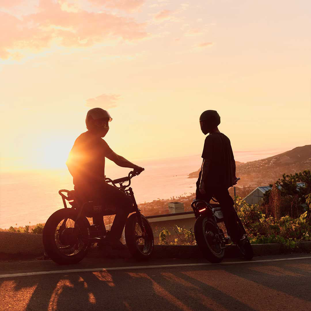 Two riders in helmets with the SUPER73 x Oracle Red Bull Racing M!D SE ebikes standing on the side of a roadway overlooking the beach at sunset.