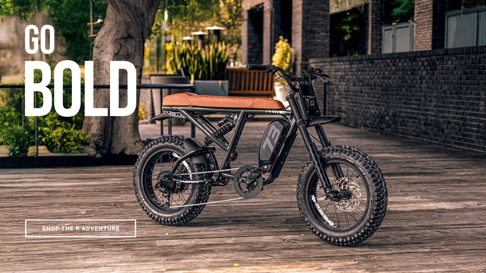 Lifestyle image of the SUPER73-R Adventure SE ebike in Palladium. The bike is parked on a wooden surface with a brick building and plants in the background.