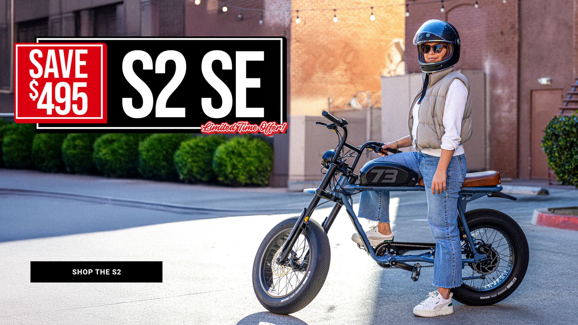 Lifestyle image of a woman wearing a helmet sitting on a SUPER73-S2 SE ebike in Panthro Blue. A text overlay on the image says "Save $495 on the S2 SE for a limited time."