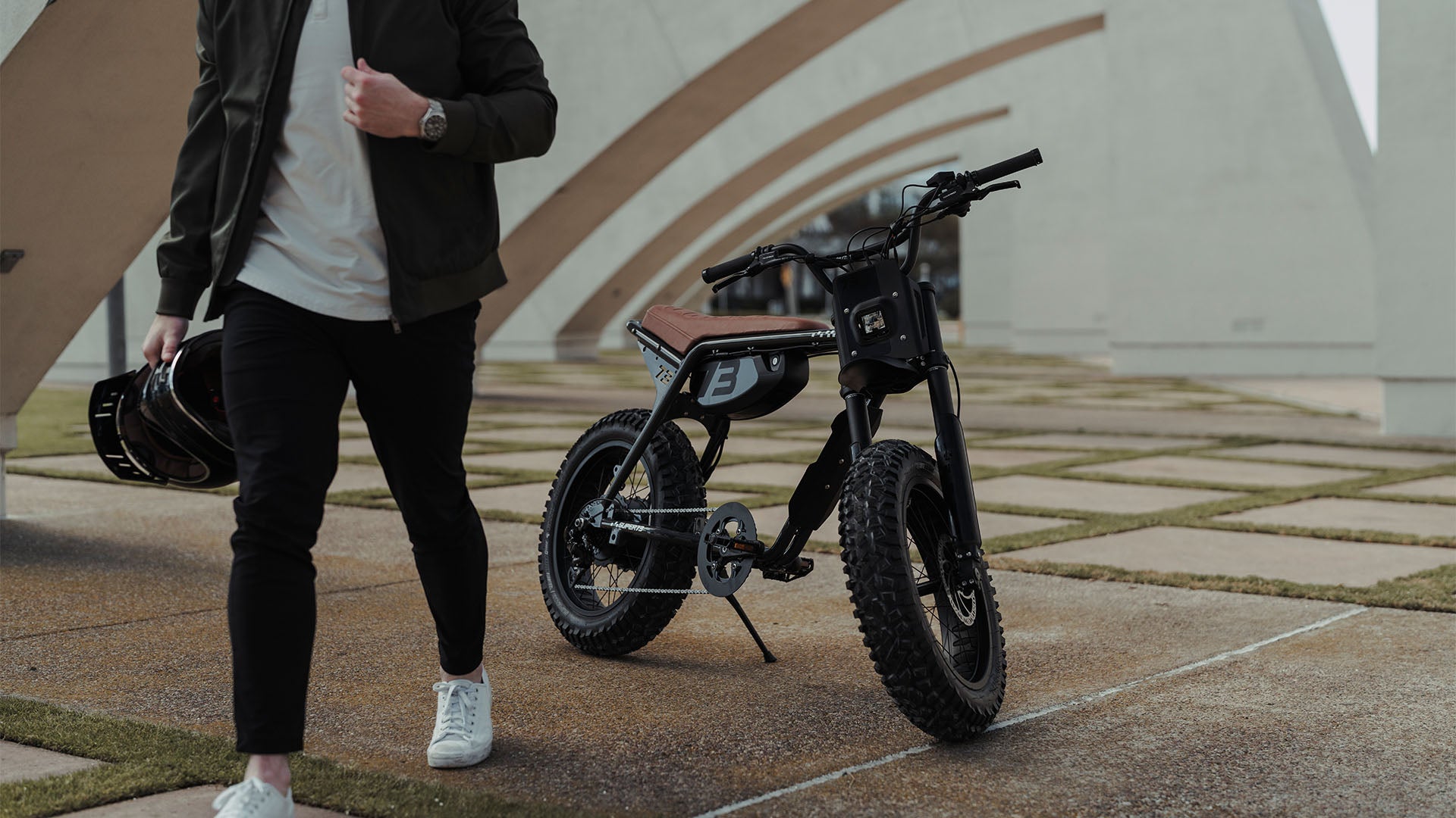 Lifestyle image of a man carrying a helmet with a SUPER73-ZX SE ebike in Bandit in the background